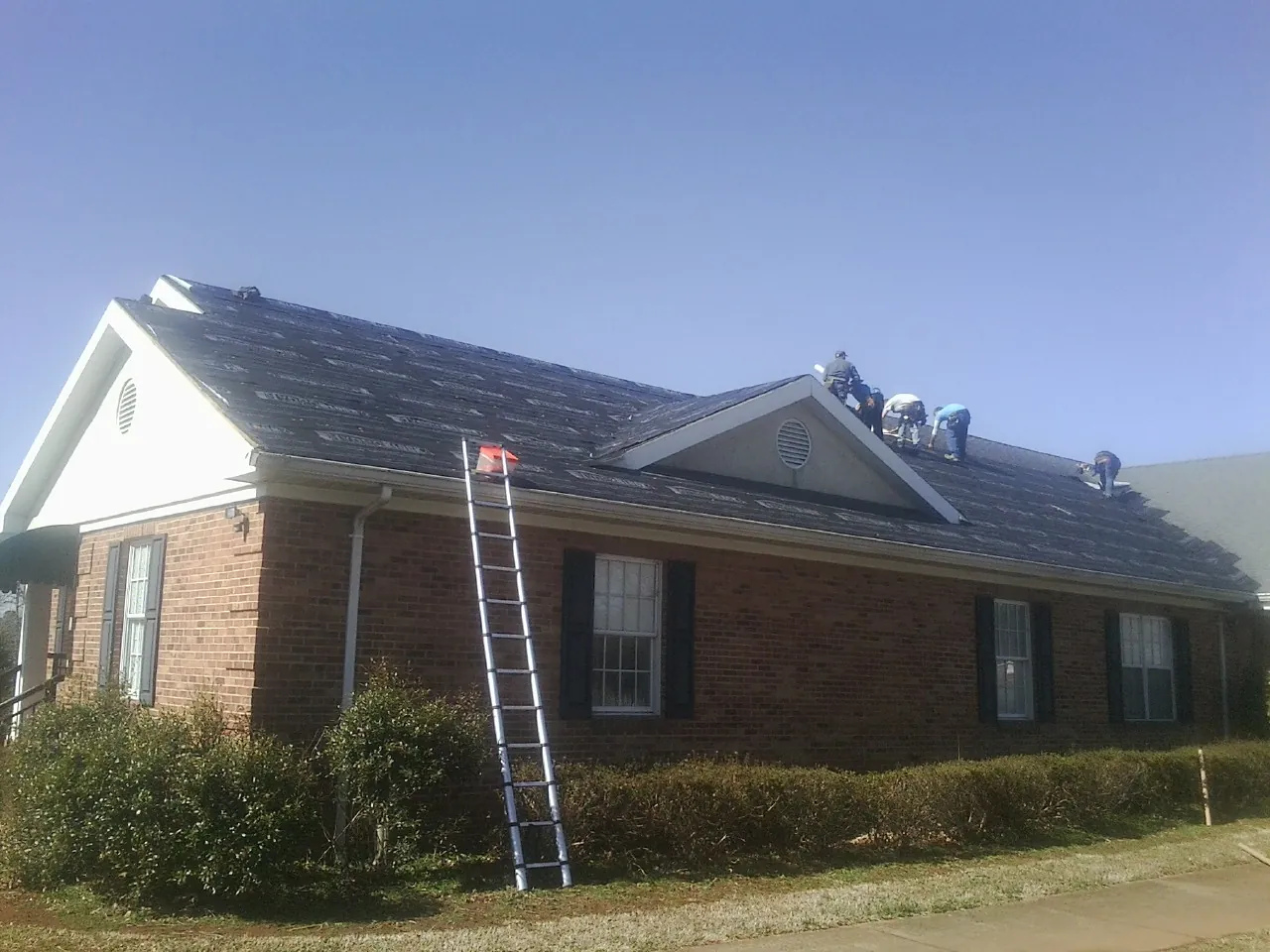 Crew tearing shingles off a brick ranch roof with a ladder set — Emergency Roof Repair work by our crew in Hague