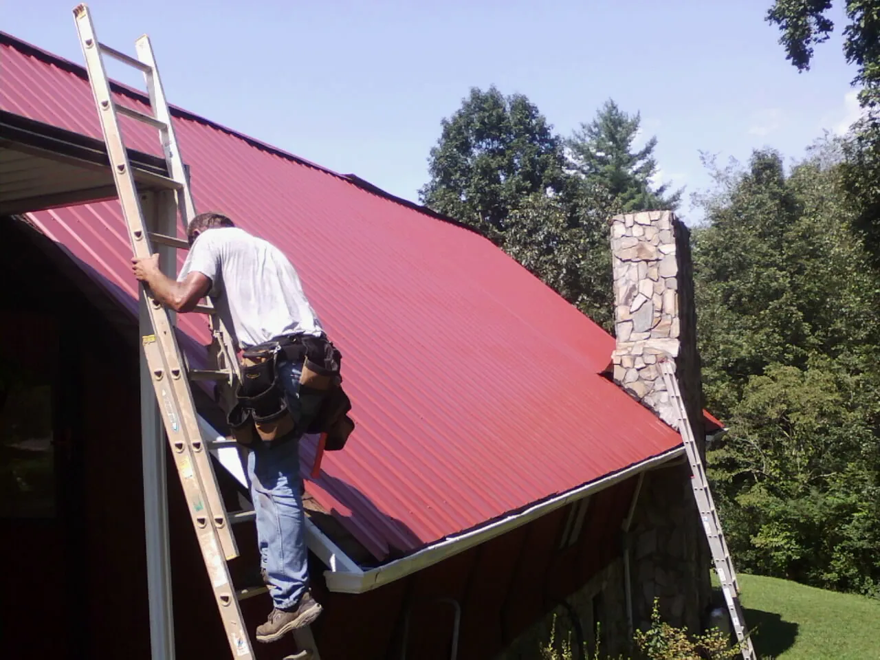 Roofer on a ladder beside a red metal roof and stone chimney — Soffit & Fascia Repair by Roofing Brothers in Hague